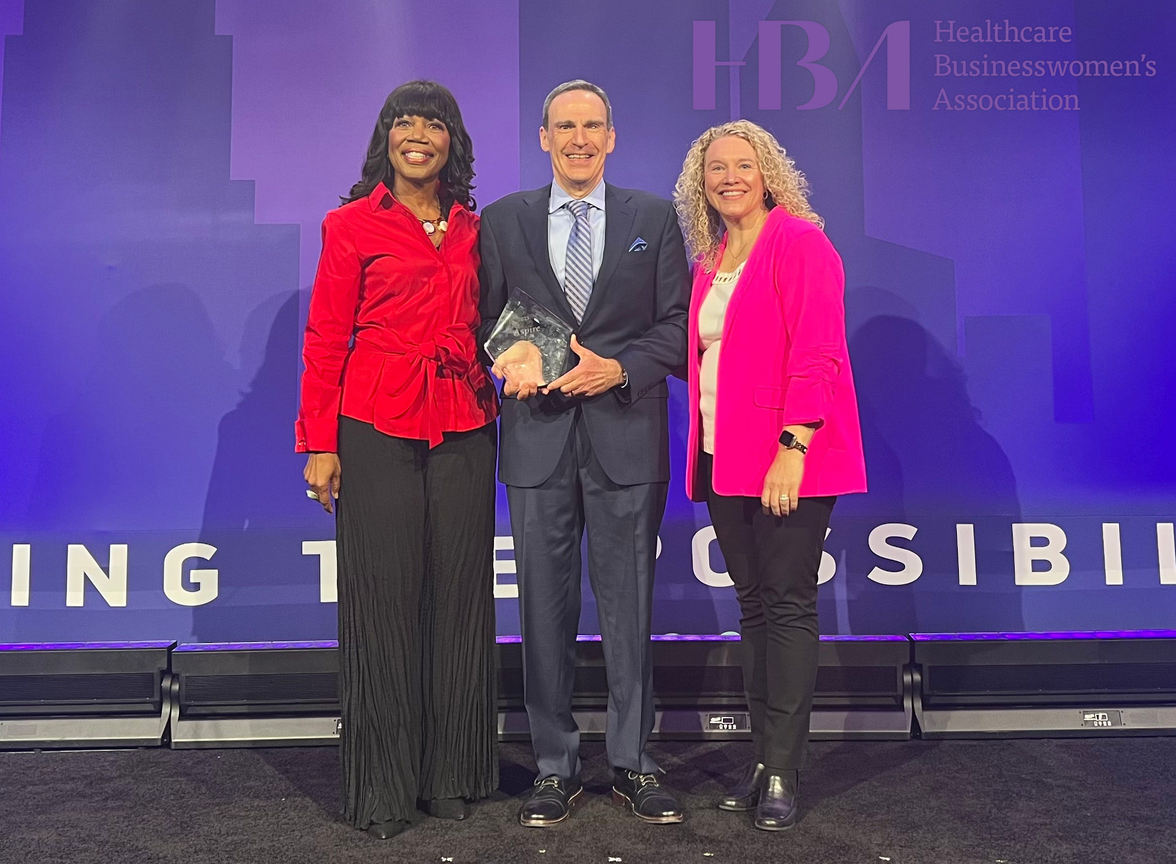 Three people on a stage. Gentleman in the middle of two women, he is holding an award.